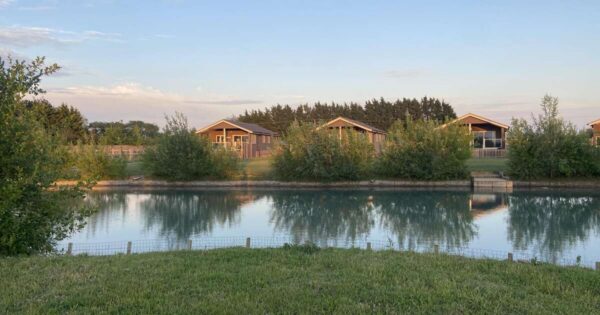 looking over a grass bank and a lake to 3 wooden lodge cabins on the far bank