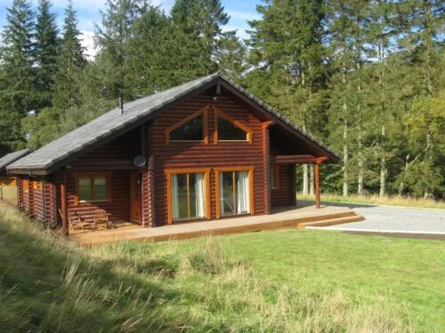 Log cabin style lodge with a veranda beside a brown trout lake, backed by conifer woodland
