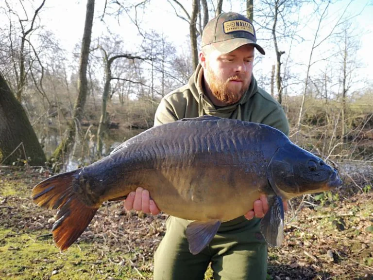 An angler holding a mirror carp beside a tree lined lake