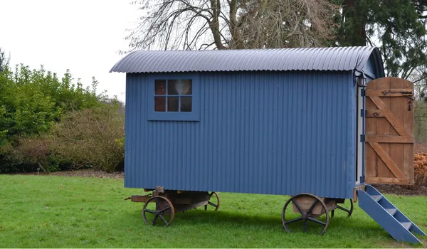Blue metal shepherds hut with cast iron wheels and wooden door on a grassy lawn