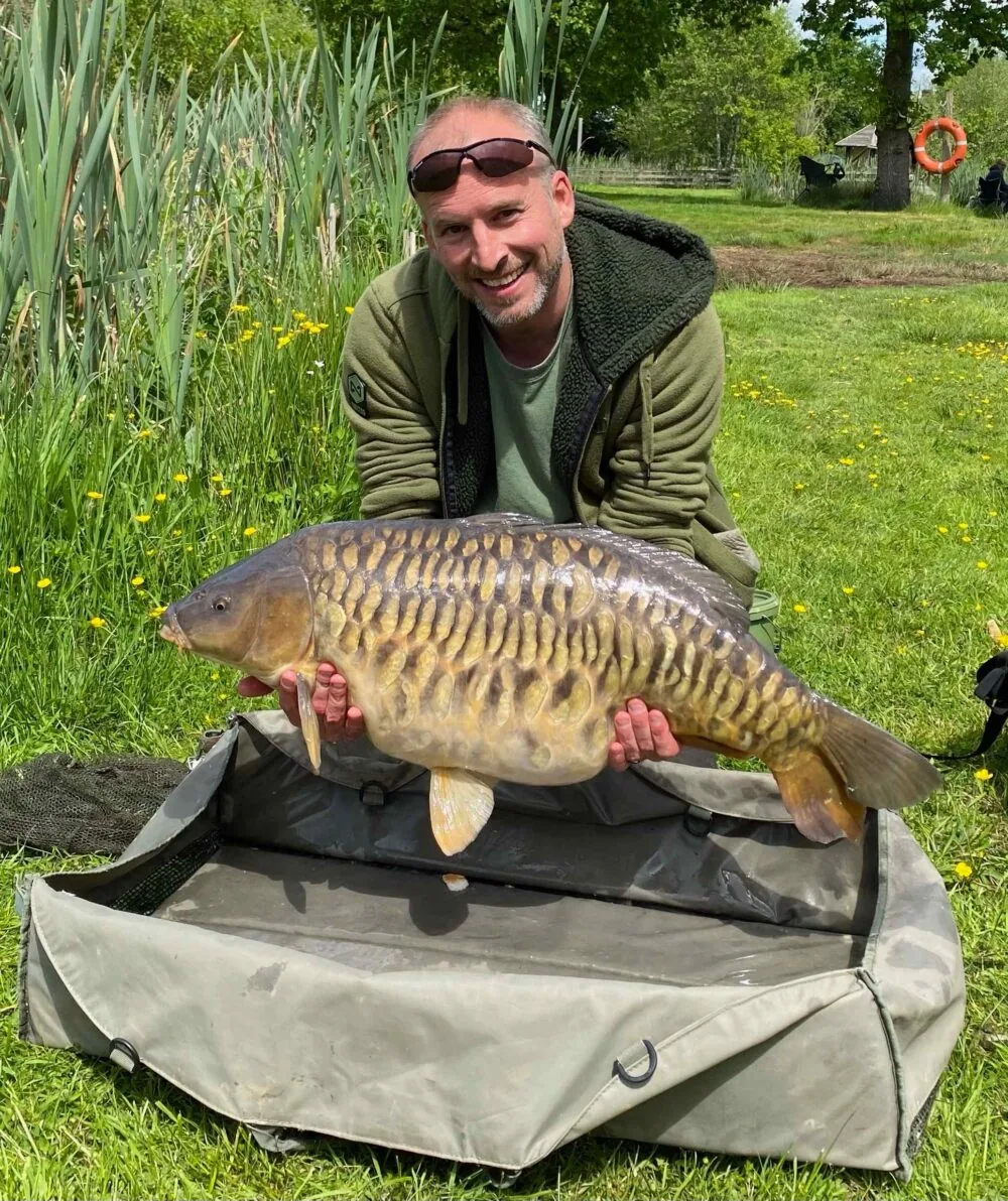 An angler kneeling on the bank holding a fully scaled carp at Spring Heath Fisheries, Shropshire