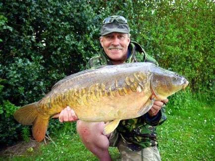 An angler holding a large mirror carp beside the water at Stenhill Private Fishing Lake, Devon