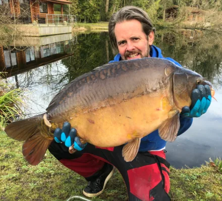 An angler holding a large mirror carp beside a lake with a timber lodge behind